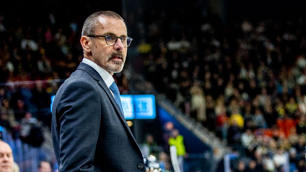 Tommy Samuelsson står iklädd kostym vid sargen under en ishockeymatch inför fullsatta läktare.