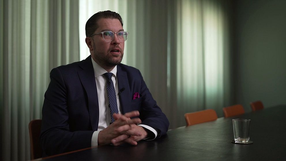 Synkbild på Jimmie Åkesson i intervju med SVT. Åkesson är klädd i kostym och sitter med händerna knäppta vid ett stort bord i ett konferensrum med fördragna garginer i dovt ljus.