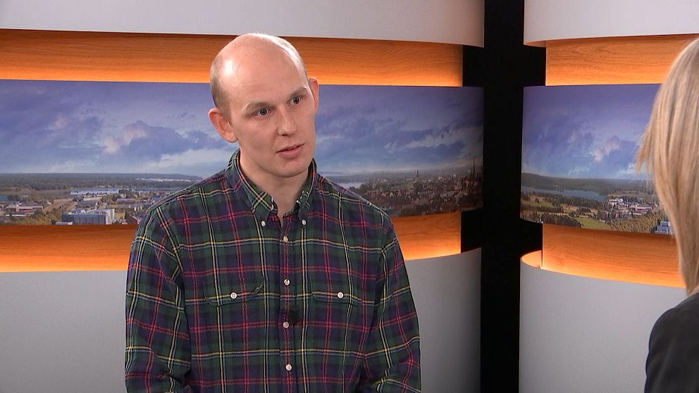 En man står i en intervju i en studio, klädd i en rutig skjorta. I bakgrunden syns panoramabilder av en stad med grönska och vatten. Belysningen är mjuk och ger en professionell atmosfär.