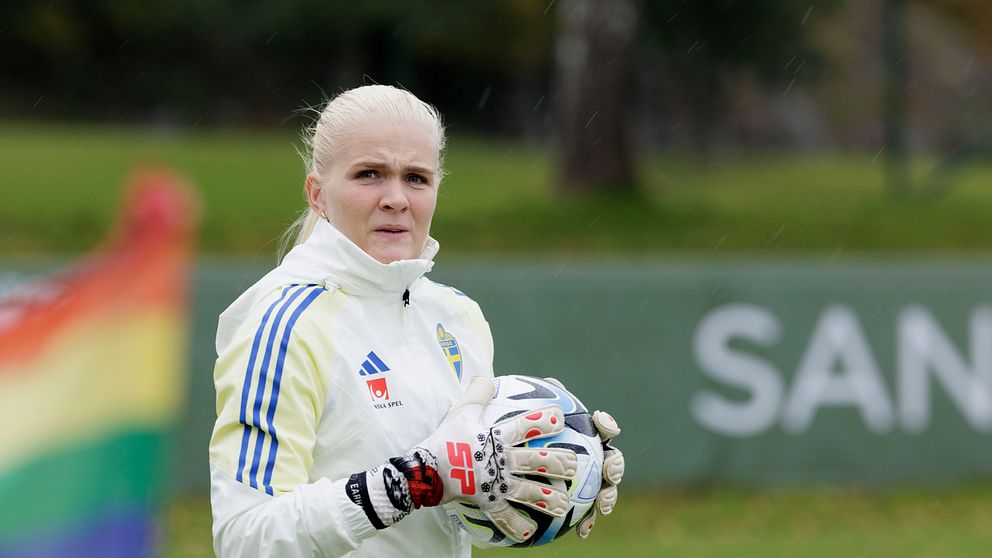 En lagmedlem i Sveriges damlandslag i fotboll står på en plan i vit och blå träningsjacka, håller en fotboll i händerna och en regnbågsflagga syns i förgrunden.