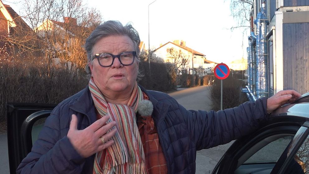 En person står brevid en bil och gestikulerar med handen, iklädd en blå jacka och en randig halsduk, mot en bakgrund av bostadshus och en vägskylt.
