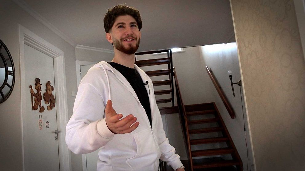 Alexander Bisso i vit hoodie står i hallen framför trappa, räcker ut handen mot kameran.