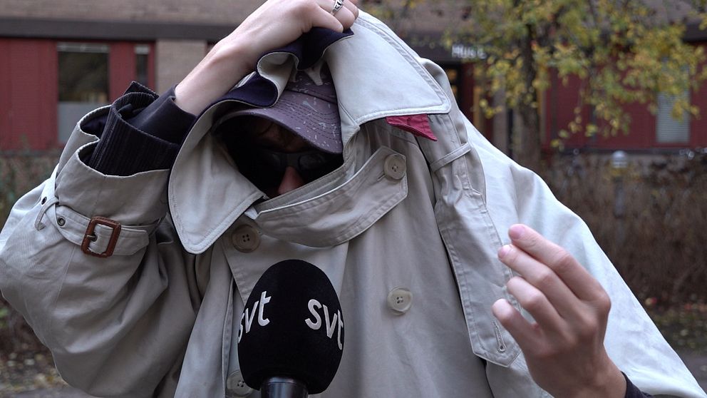En person i en beige trench coat håller upp kappan framför sig medan de talar. En mikrofon med logotypen för SVT syns i nederkanten av bilden. Bakgrunden visar en del av en byggnad med röda fönster och omgivande natur med höstlöv.