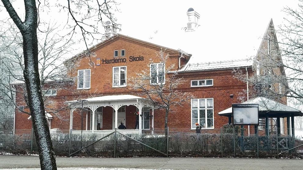 Röd tegelbyggnad som utgör Hardemo Skola, omgiven av snötäckta träd och en vinterlandskap. Byggnaden har en traditionell stil med en veranda och flera fönster. En del av skolan är annoterad med skolans namn.