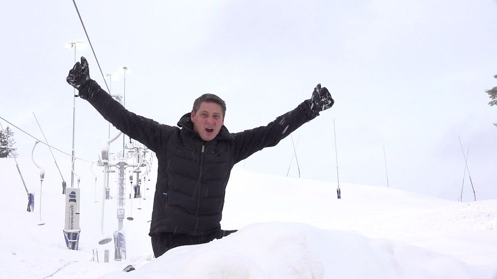 En glad man sitter i en snöig skidbacke framför en skidlift