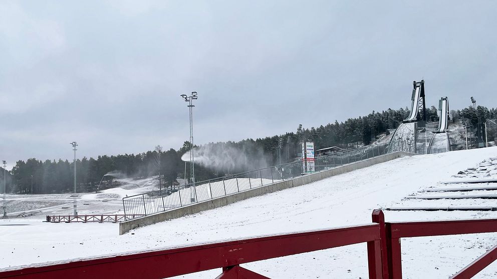 Skidstadion på Lugnet med snö och snökanoner under en kall och molnig dag. I bakgrunden syns skidspår och hoppbackar, medan ett röd räcke är synligt i förgrunden.