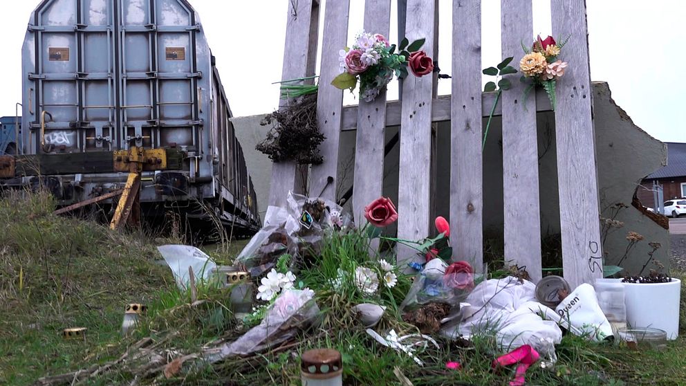 En enkel bild av en tillfällig minnesplats vid en järnvägskorsning. En träplankvägg som är dekorerad med konstgjorda blommor står i förgrunden, omgiven av skräp och sörjande föremål som ljus och blommor. I bakgrunden syns en tågvagn.