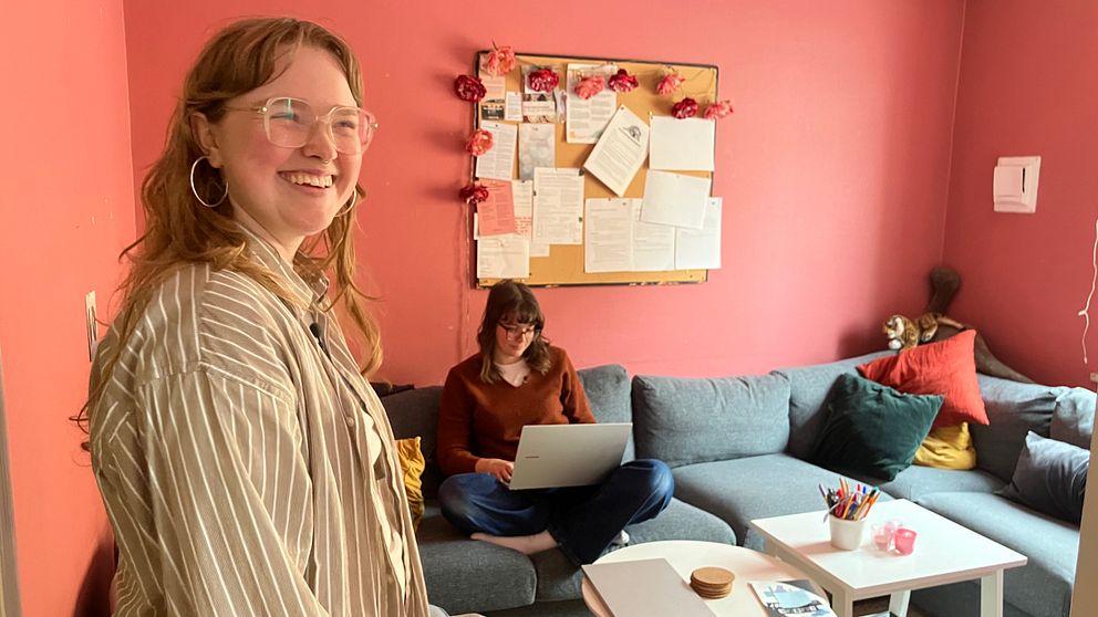 Två personer i ett färgglatt vardagsrum, en sitter i soffan med en dator och en står vid sidan.