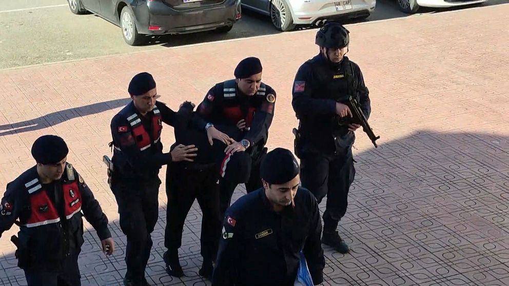 Flera poliser i uniform eskorterar en person i handfängsel. En av poliserna håller ett automatvapen. Scenen utspelar sig utomhus på en trottoar vid parkerade bilar.