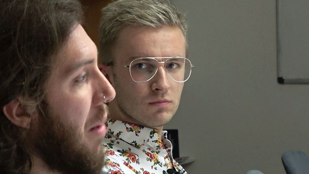 Två personer sitter nära varandra, delvis vända mot bildens vänstra sida. Den ena har lockigt hår och den andra har rakt hår. Båda bär kläder med mönster, och de befinner sig i en inomhusmiljö med hyllor i bakgrunden.