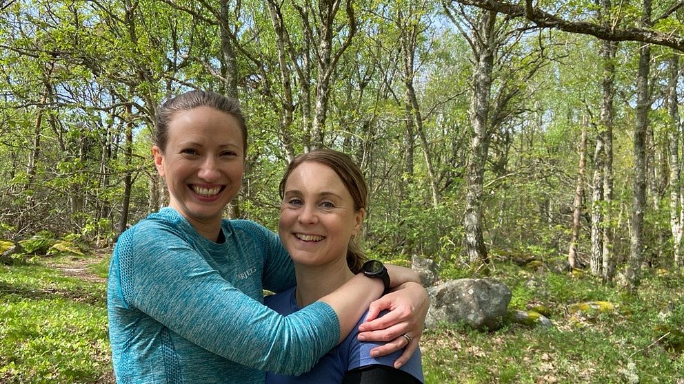 Två kvinnor i en skog kramar om varandra och ler.