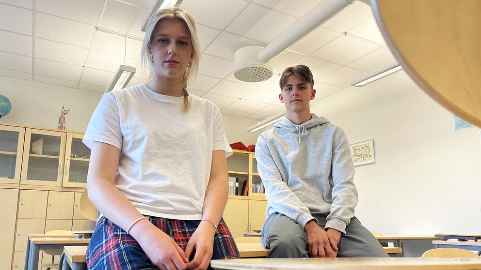 En blond kille och tjej sitter bredvid varandra på en skolbänk i ett klassrum och kollar in i kameran.