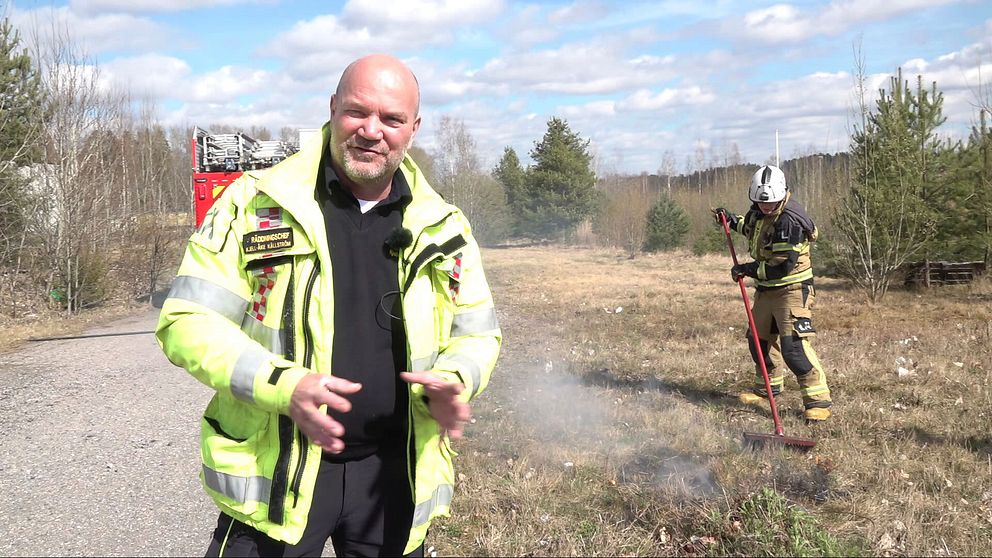 Brandman släcker mindre gräsbrand nära väg, brandbil i bakgrunden.