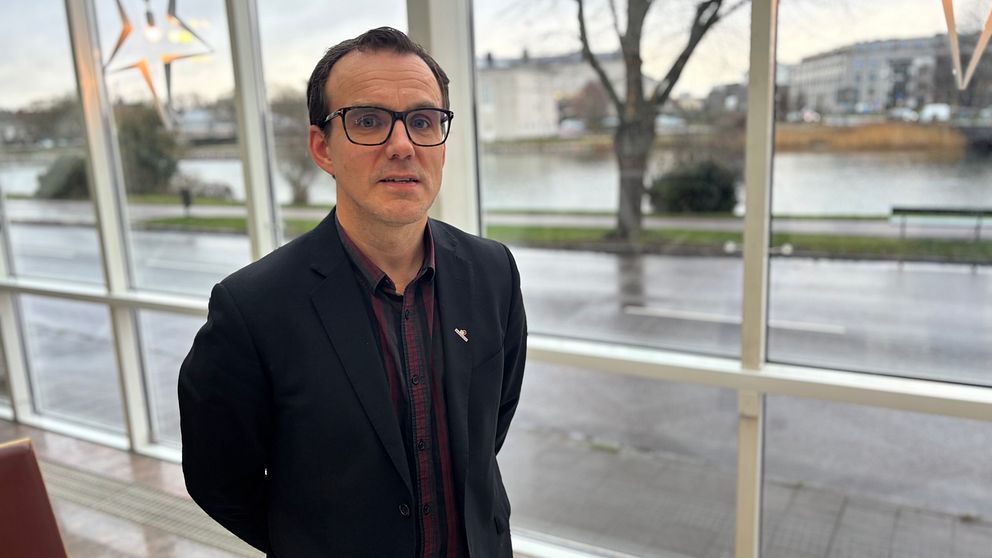 En person står framför ett stort fönster med utsikt över en flod och en stadsmiljö. Det dämpade ljuset inomhus kontrasterar med den ljusare omgivningen utanför. Det är en modern inredning med stiliga detaljer.