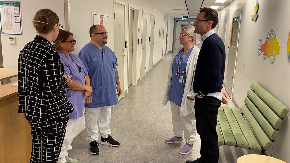 Grupp av vårdpersonal på Sundsvalls sjukhus engagerad i samtal. Två personer i sjukhusuniformer står mitt i korridoren, medan en person i kostym står i samtal med dem. En tredje person i sjukhuskläder lyssnar. Bakgrunden visar en ljus och modern sjukhuskorridor med dörrar och en sittgrupp.