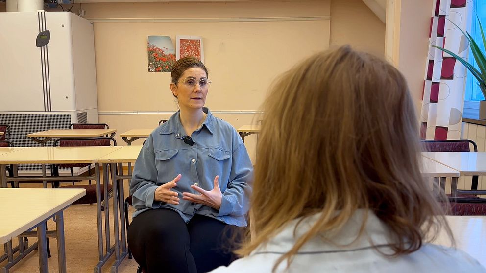 Två personer sitter vid skolbänkar och har ett samtal i ett klassrum.