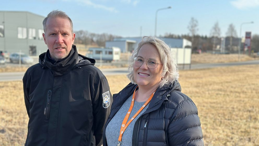 Två personer står på en gräsfält med byggnader i bakgrunden.