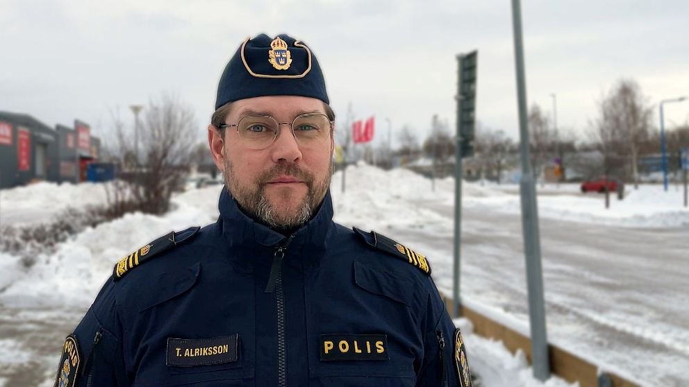 En polis i uniform står i ett snöigt stadsområde med en väg och flaggor i bakgrunden.