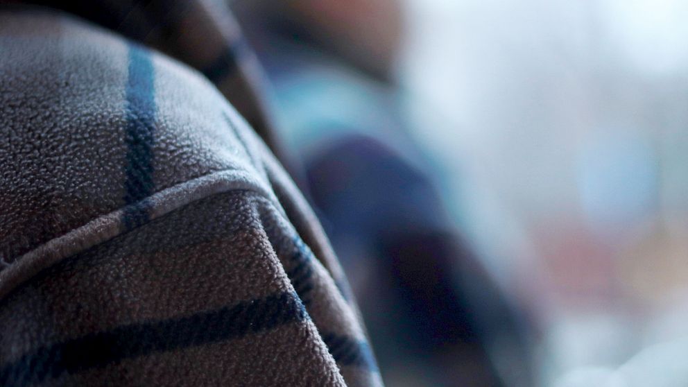 Närbild av en persons axel iklädd en rutig fleecejacka med fokus på tygets textur medan bakgrunden är oskarp och visar vaga konturer av ytterligare personer eller föremål.