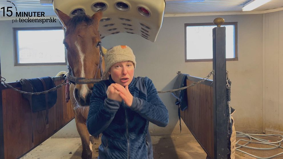 Jenny Smideblad står i ett stall och tecknar ordet stöd. Bakom henne står en häst i en spilta.