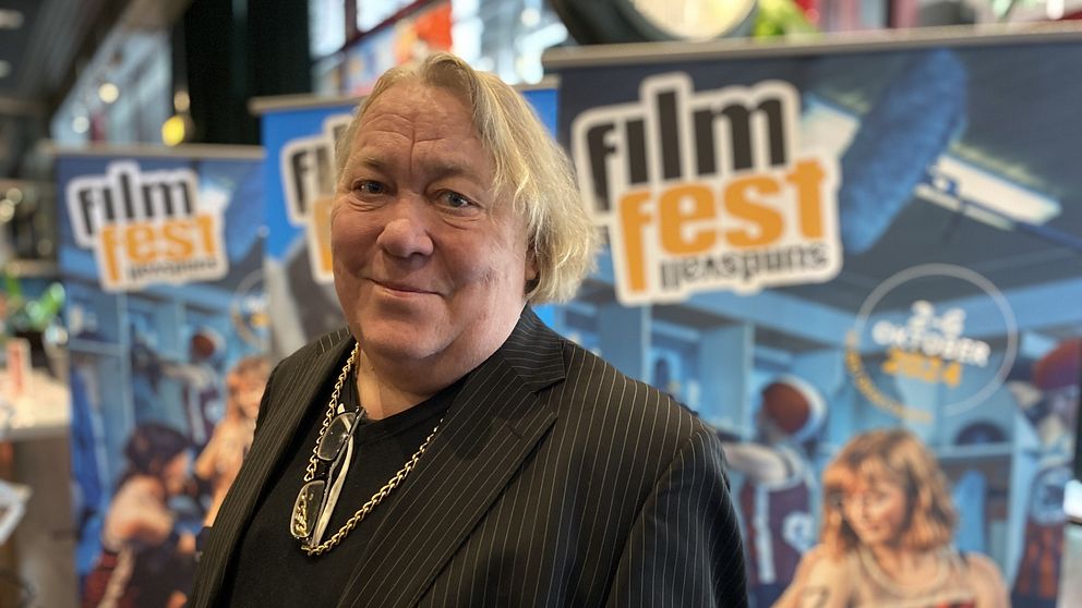 Leif Andrée iklädd en svart randig kavaj står framför en bakgrund med affischer för ”Filmfest” som visar olika scener från filmfestivalen. Affischerna har färgglada bilder av filmiska element och texten ”film fest”. Miljön ser ut att vara ett evenemangsutrymme.