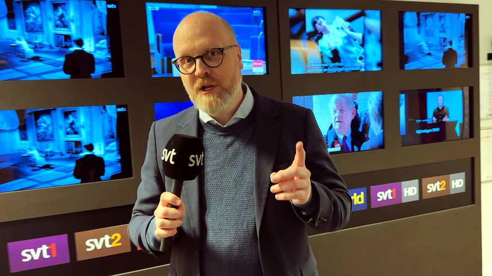 En person håller en mikrofon med SVT-logga framför en vägg av TV-skärmar.