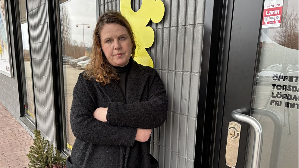 En person med axellångt hår står framför en butiksentré med glasdörrar, klädd i en svart jacka. I bakgrunden syns en del av en gul skylt och öppettiderna för butiken.