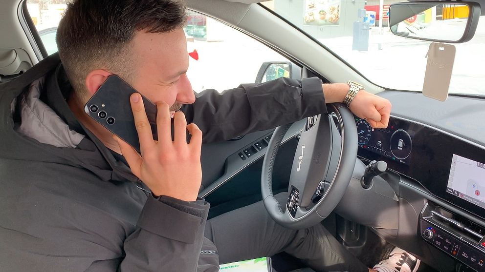En man sitter i en bil och pratar i telefon, andra handen har han på ratten.