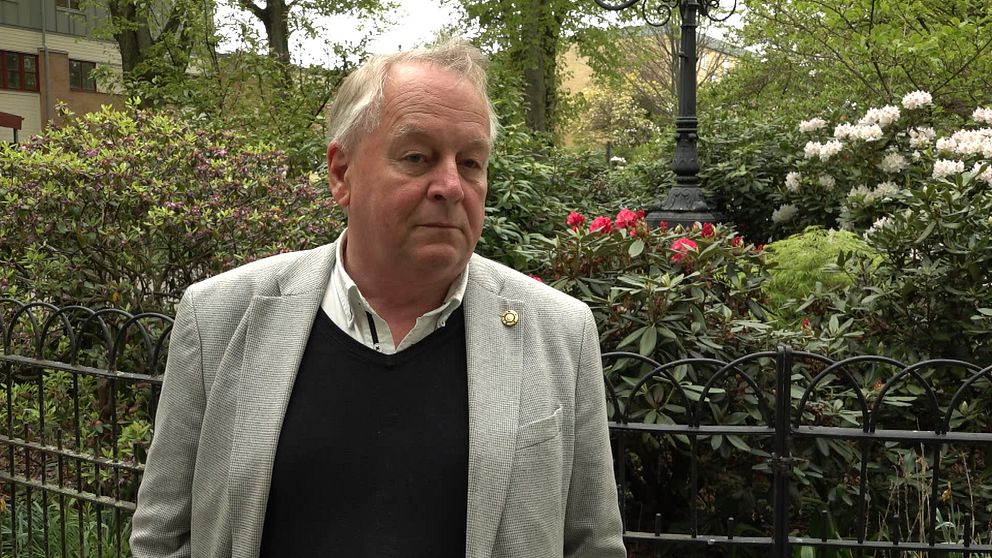 Sven Andersson i grå kavaj står vid ett svart staket i grönskande parkmiljö.