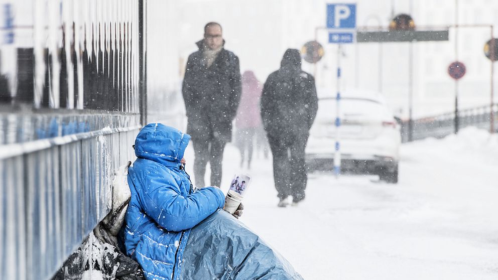 EU-migrant sitter och tigger, har vinterkläder på sig och det snöar.