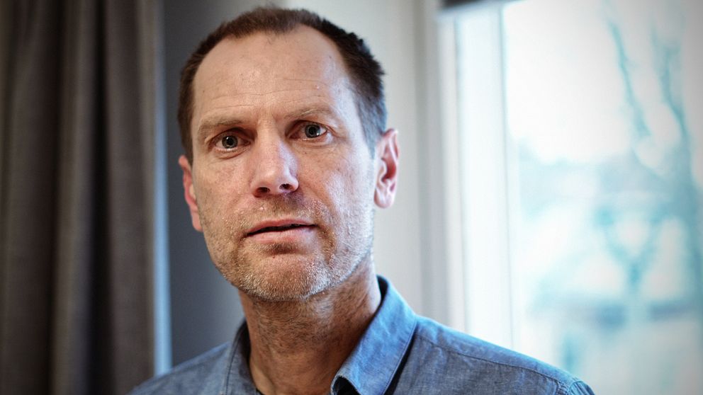 En person står inomhus nära ett fönster iklädd en jeansskjorta, med gardiner synliga i bakgrunden.