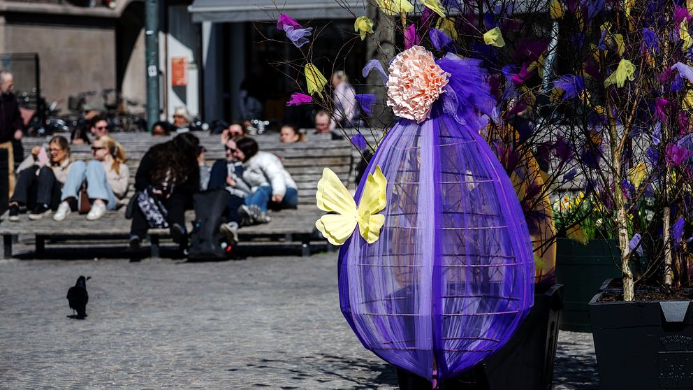 Utsmyckat påskträd på torg. Människor sitter på bänkar i bakgrunden.