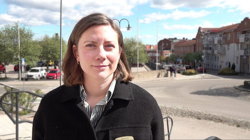 Person med axellångt hår står utomhus vid ett torg med byggnader och träd i bakgrunden.