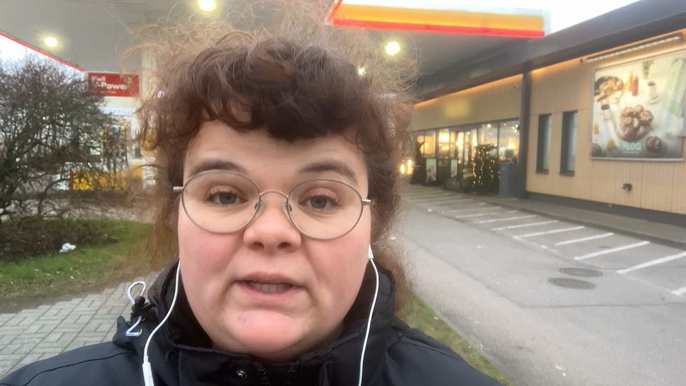 Reporter Klara Misgeld står utomhus i en stadsmiljö nära en bensinstation, med en byggnad i bakgrunden som tyder på kommersiell verksamhet. Det är en grå och mulen dag. Personen har hörlurar och bär en svart jacka. Det finns parkering i bakgrunden tillsammans med buskar och en del av en gångväg.