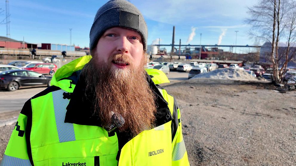 Tomas Hart med lysande grön jacka står vid industriområde med bilar och fabriker i bakgrunden.