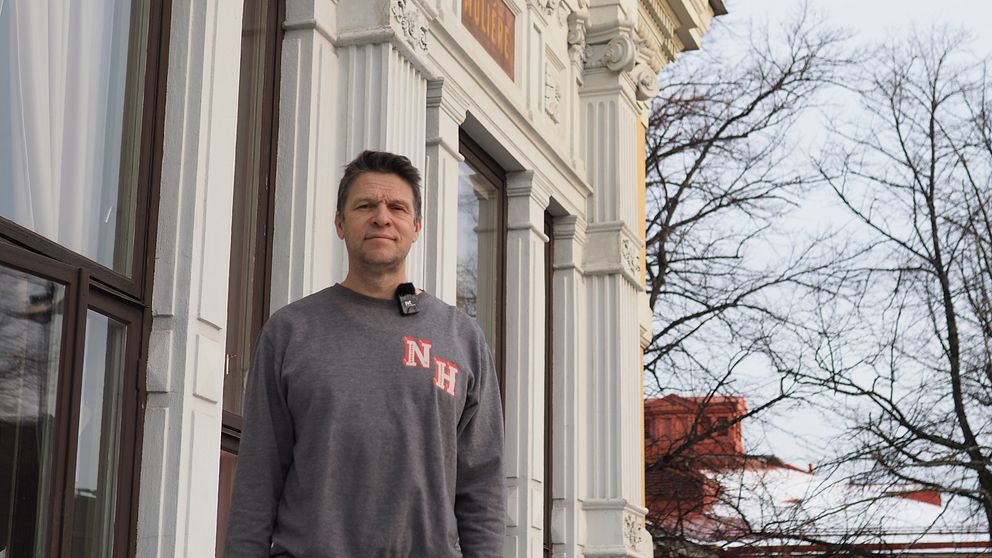 En person står utanför en byggnad med klassisk arkitektur, inklusive dekorerade fönster och detaljerad fasad. Bakgrunden visar träd utan löv, vilket ger en vinterliknande atmosfär. Personen bär en gråtröja med ett rött mönster på bröstet.