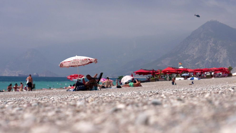 Turister vid populär strand med parasoller och berg i bakgrunden.