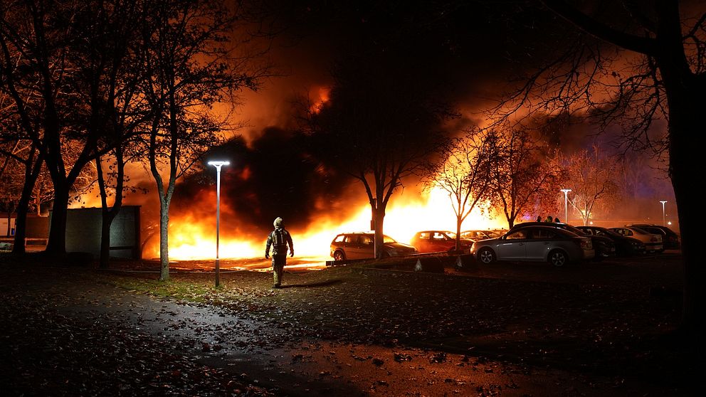 En person går genom en parkering i skymningen, med en stor brand som rasar i bakgrunden. Flammor och rök lyser upp natten, medan träden och bilarna ger en kontrast till den dramatiska scenen.