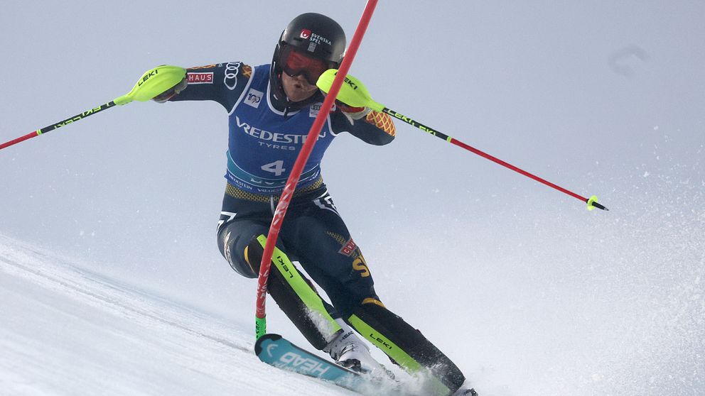 En alpin åkare i färgglada skidkläder åker nerför en snöiga backe, med fokus på teknik och hastighet. Åkaren använder skistavar för balans medan snön sprutar upp runt dem.