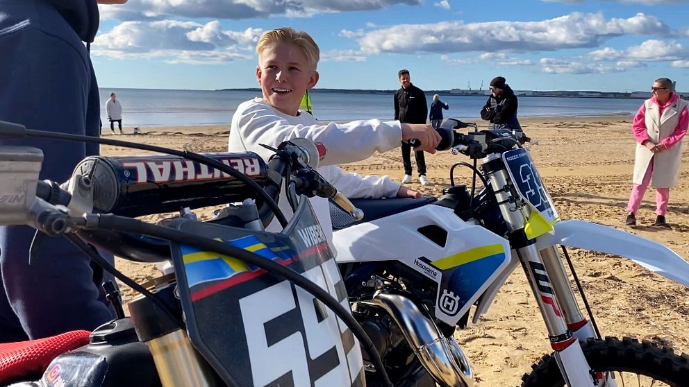 En pojke sitter på en motorcrosscykel på en sandstrand, omgiven av flera personer som står i bakgrunden. Det är en solig dag med några moln på himlen och havet syns i fjärran. Cykeln i fokus är vit med blå och gula detaljer, medan en annan motorcykel med nummer 54 också syns i bildens främre del.