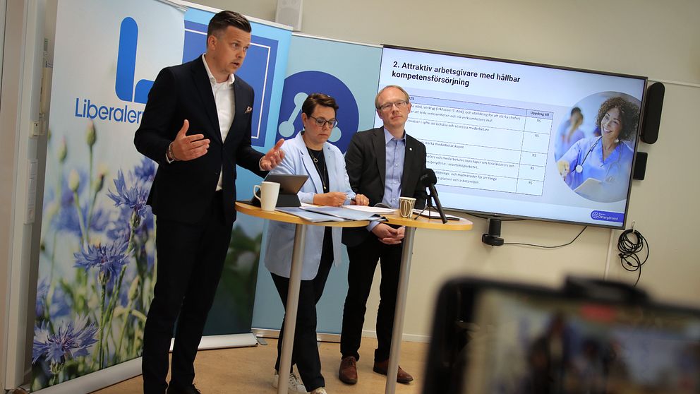 Tre politiker står framför en skärm i ett konferensrum och håller en presentation. En av de, en man i kavaj och vit skjorta gestikulerar med händerna.