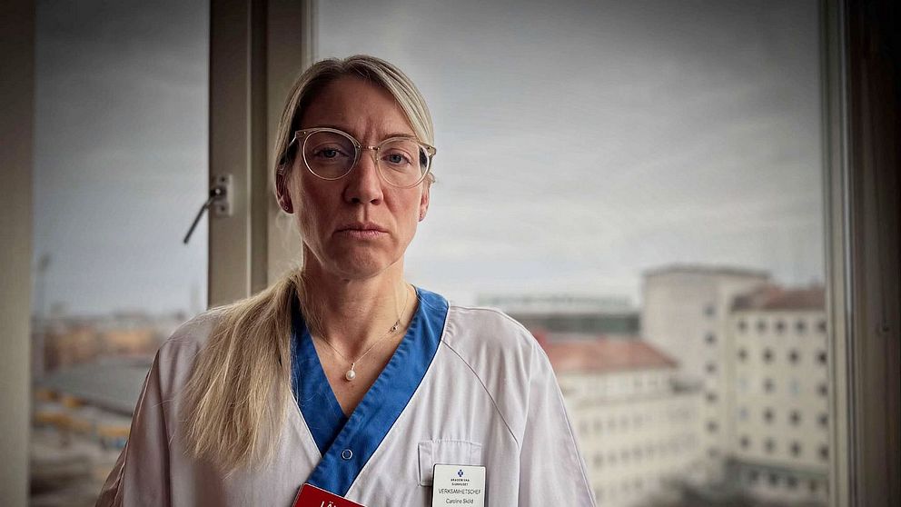 Sjukvårdspersonal i vit uniform står vid fönster med sjukhusbyggnader i bakgrunden.