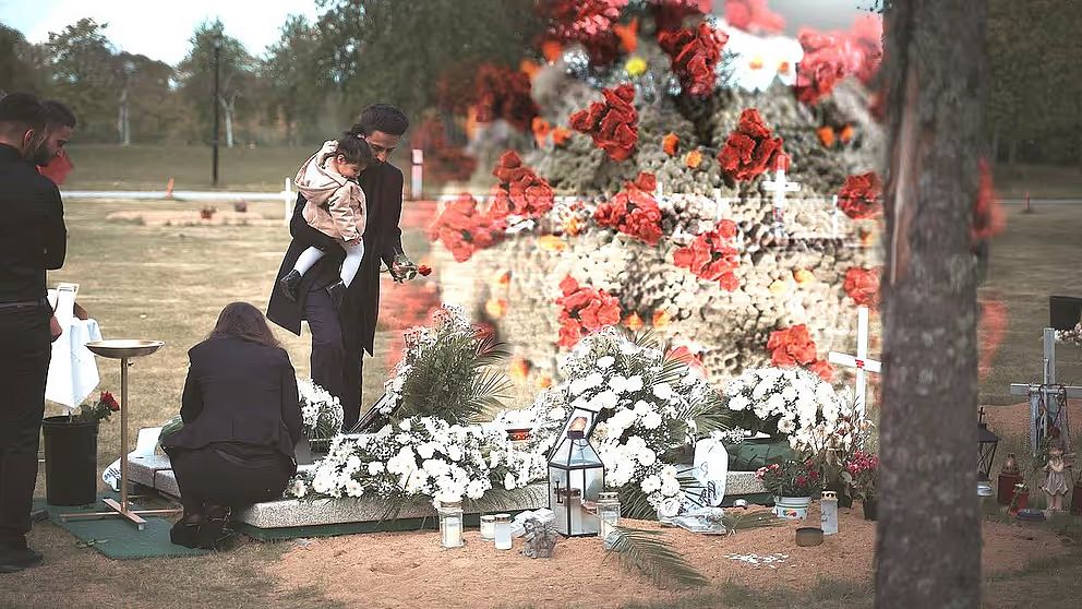 En familj samlas vid en gravplats för att hedra en avliden person. Två personer står vid graven, där en man håller ett barn, medan en kvinna sitter på knä vid gravstenen. Omkring graven finns det en stor uppsättning blommor, mestadels vita, tillsammans med ljus och andra minnesföremål. I bakgrunden syns en närbild av coronaviruset.