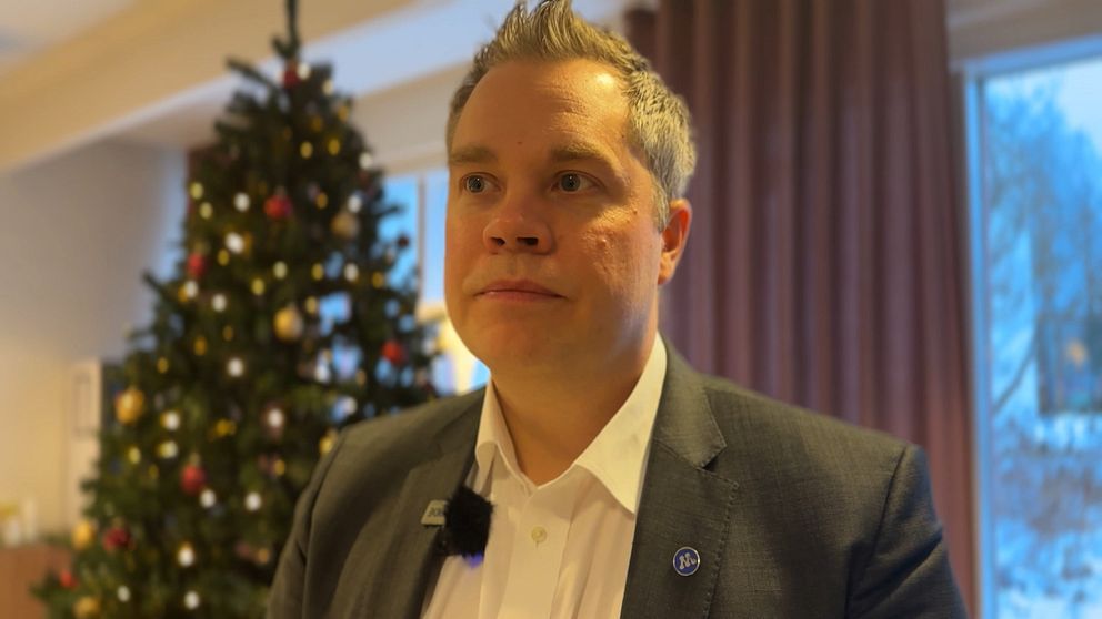 En person står framför en julgran dekorerad med färgglada kulor och ljus. Bakgrunden visar ett rum med gardiner i dämpade färger och ett fönster som släpper in naturligt ljus. Personen bär en mörk kostym med en vit skjorta och har en mikrofon fäst vid kragen.