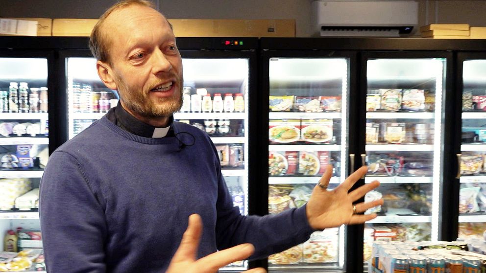 En person i en blå tröja och prästkrage gestikulerar framför ett kylskåp fyllt med matvaror och drycker i en affär eller butik.