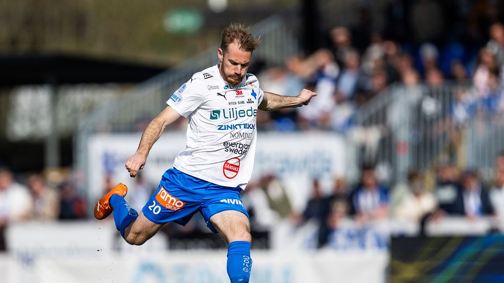 Fotbollsspelare sparkar boll under match på gräsplan med publik i bakgrunden.