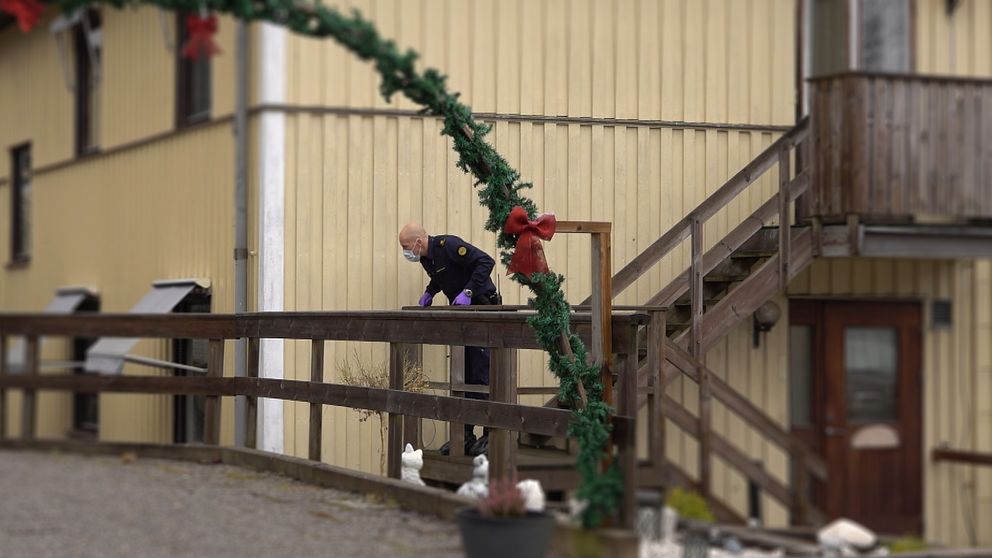 En polis med ansiktsmask och handskar undersöker något vid ett räcke utanför ett gult hus, dekorerat med grön girland och röda rosetter.