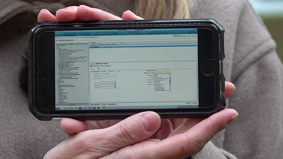 En person håller upp en smartphone som visar en skärm med programvara, med olika alternativ och fält för information. Skärmen verkar vara en del av ett gränssnitt för datainmatning eller hantering.