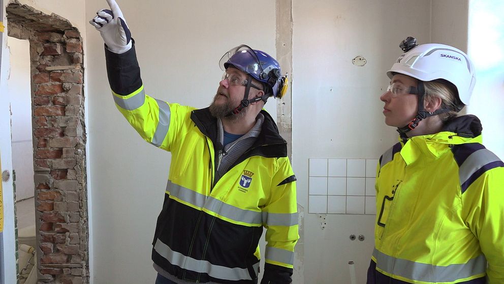 En man och en kvinna i varseljackor och bygghjälmar står och tittar upp på en vägg som håller på att renoveras inne i Rådhuset i Östersund.