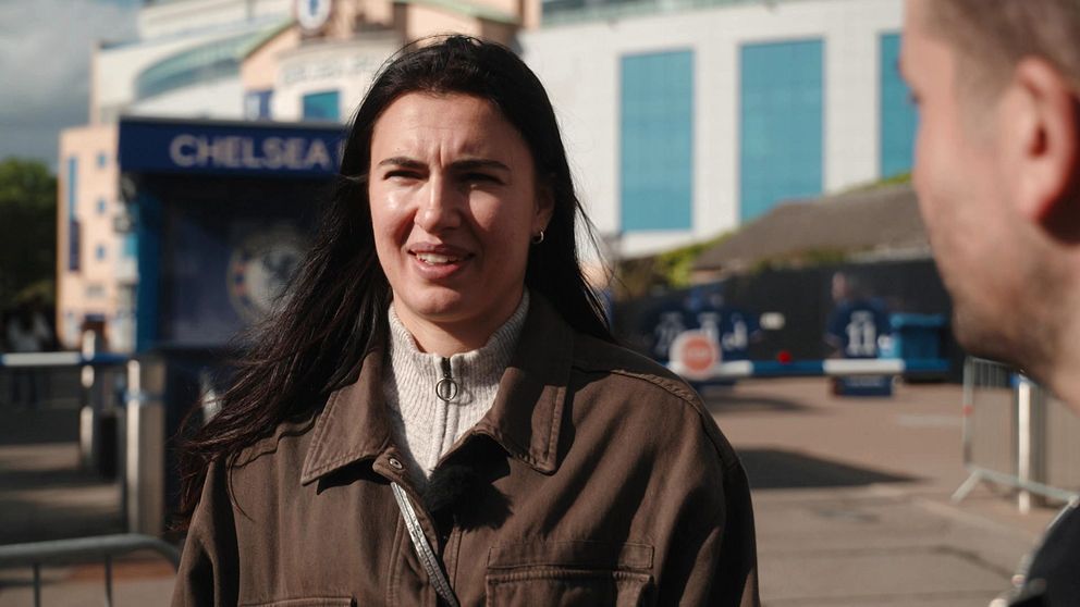 Två personer samtalar utanför Chelseas stadion Stamford Bridge i soligt väder.